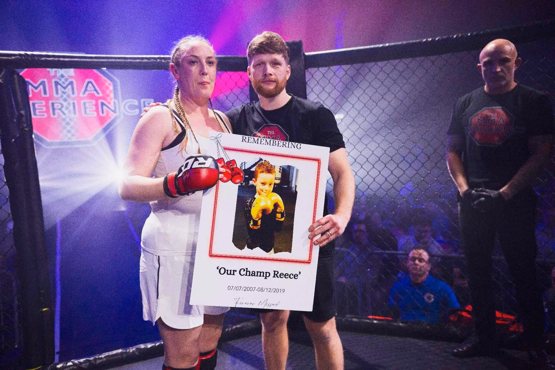 A woman in boxing clothes stands next to a man. They both hold a large banner featuring a photo of their son.