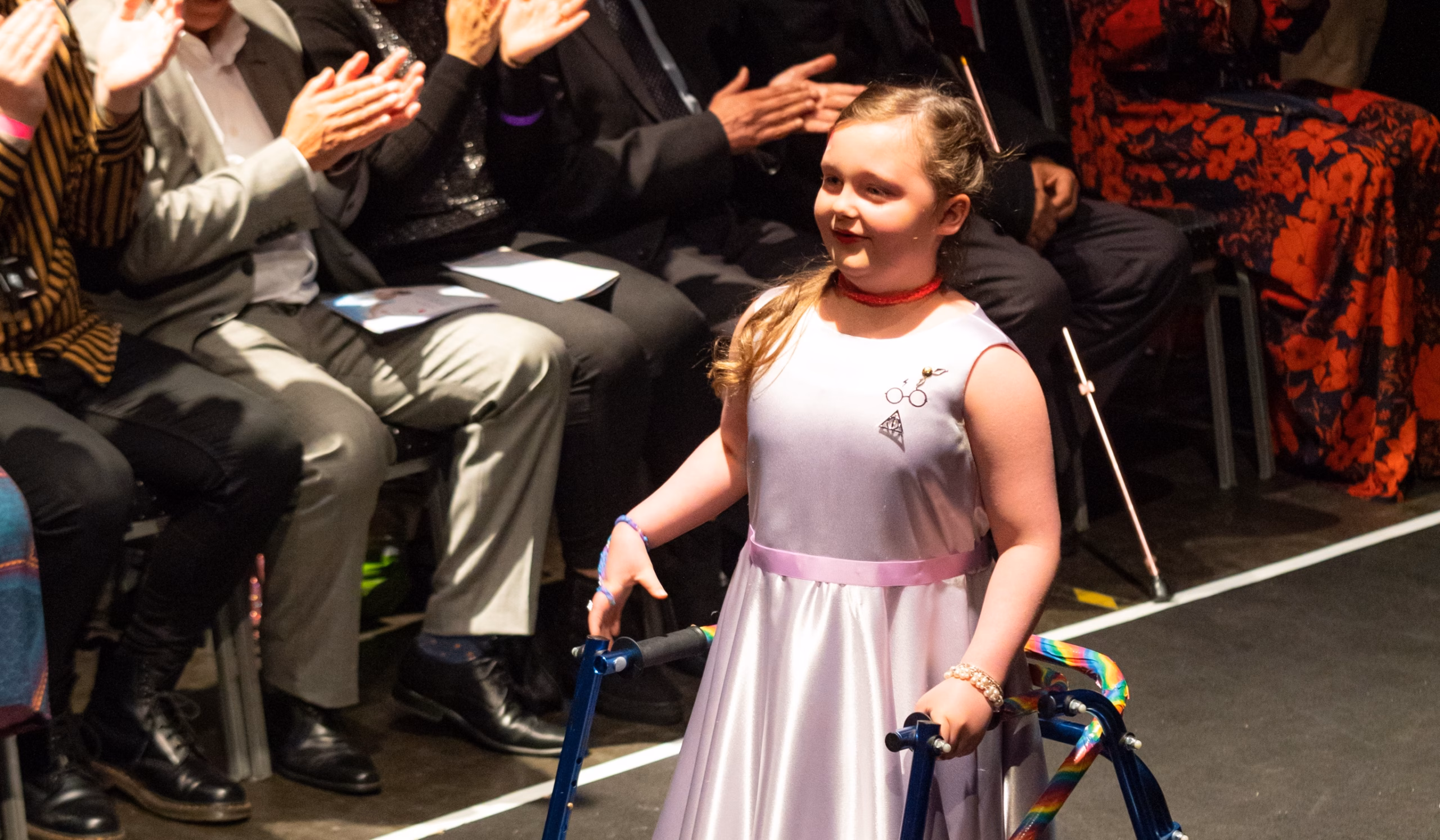 A young girl with a walking frame walks down a catwalk wearing a light pink dress