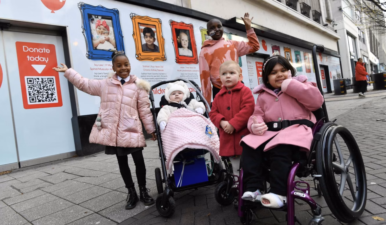 A group of children pose for a photo. Three are standing, one is in a pushchair, and one is in a wheelchair.