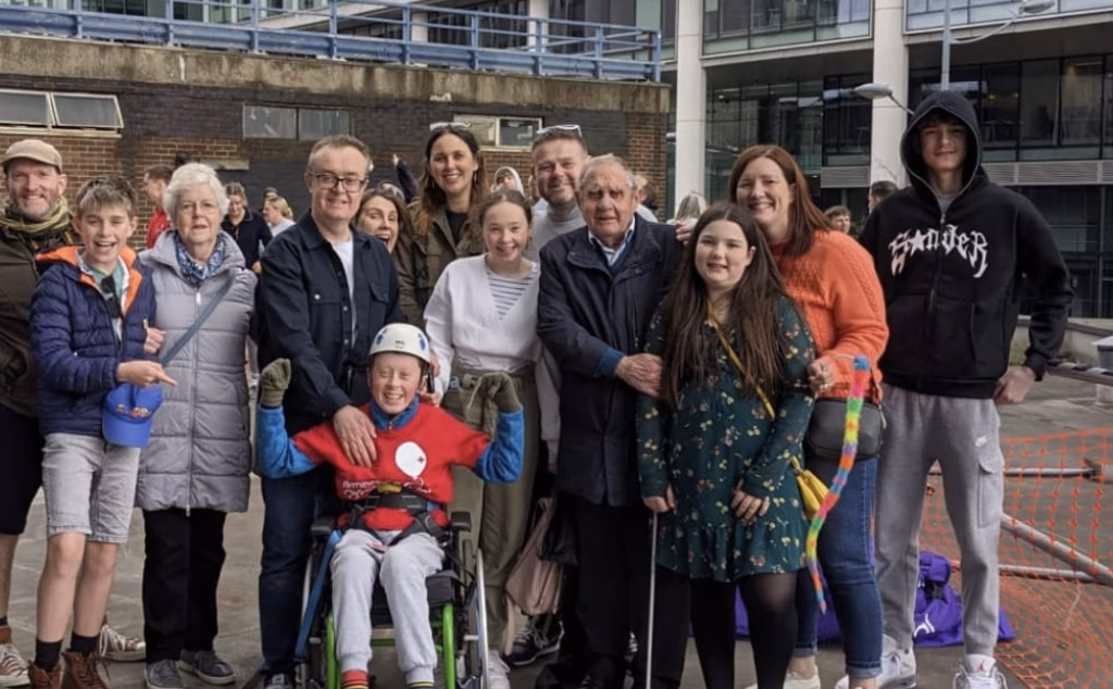 A large group of people pose together for a photo. In the middle is a child in a wheelchair wearing a red Birmingham Children's Hospital t-shirt.