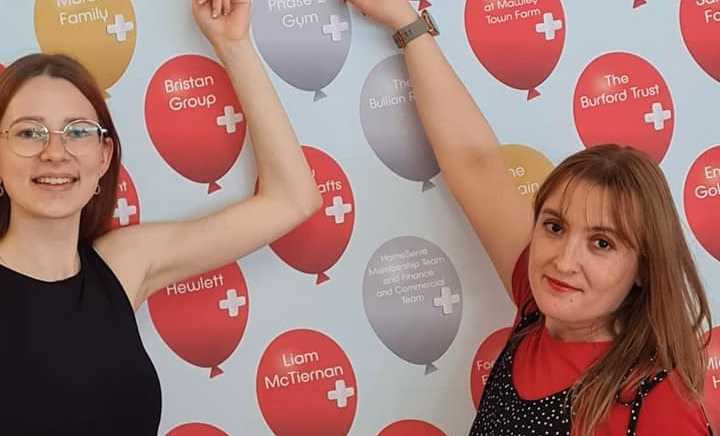 Two young women point to a step-and-repeat board featuring images of balloons. The balloon they point to has their company's name on it.