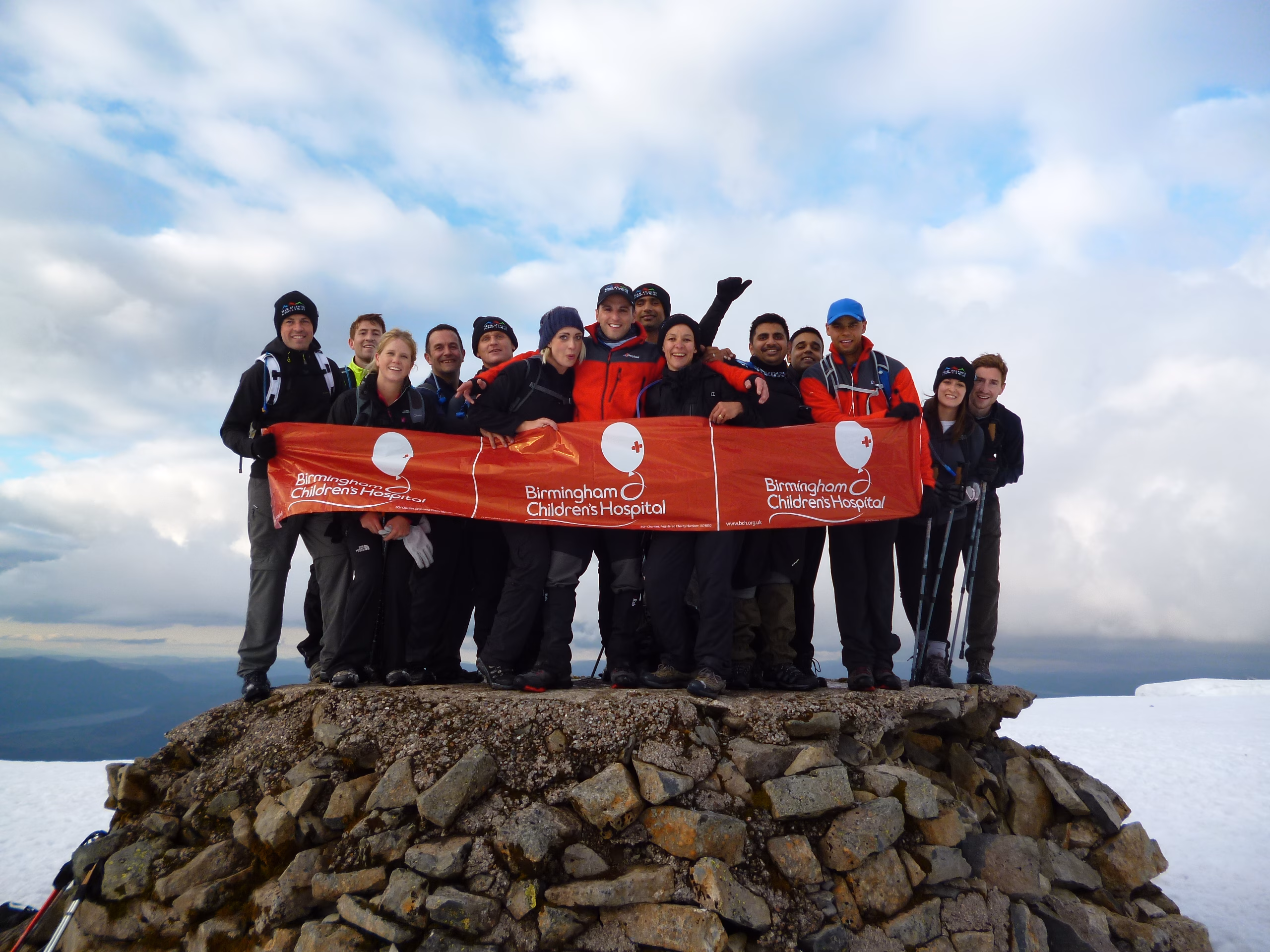 A group of men and women stand on top of a summit. They hold a large Birmingham Children's Hospital Charity banner.
