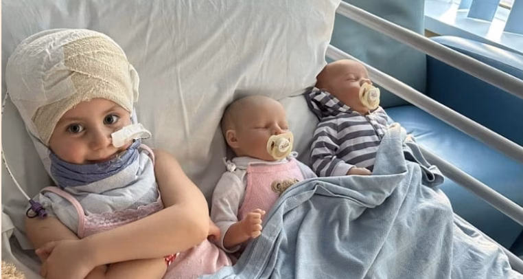 A young girl in a hospital bed with a bandage around her head. There are two toy dolls in the bed next to her.