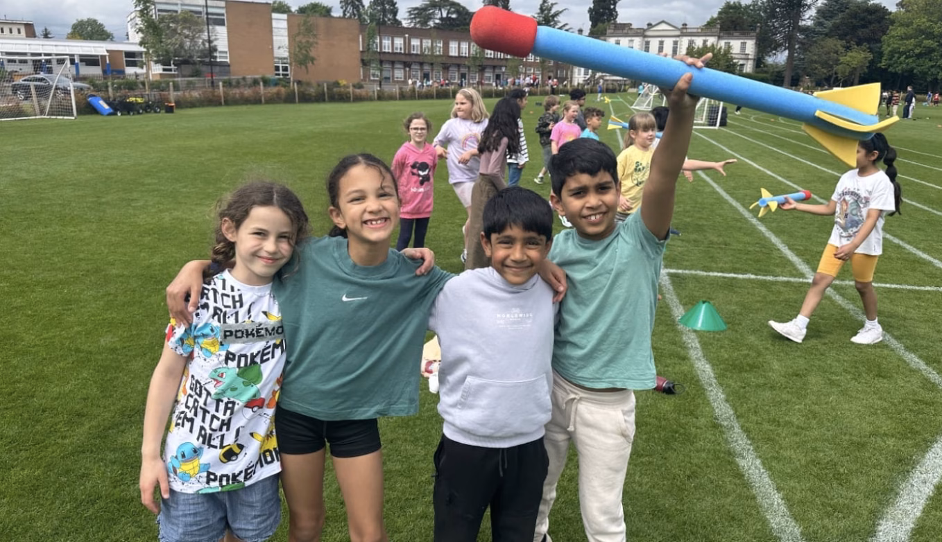 A group of children stand on a school field. One holds a foam baton above his head.