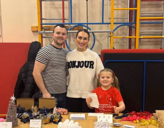 A dad, mum and little girl stand in a school gym behind a table. The table features items being sold.