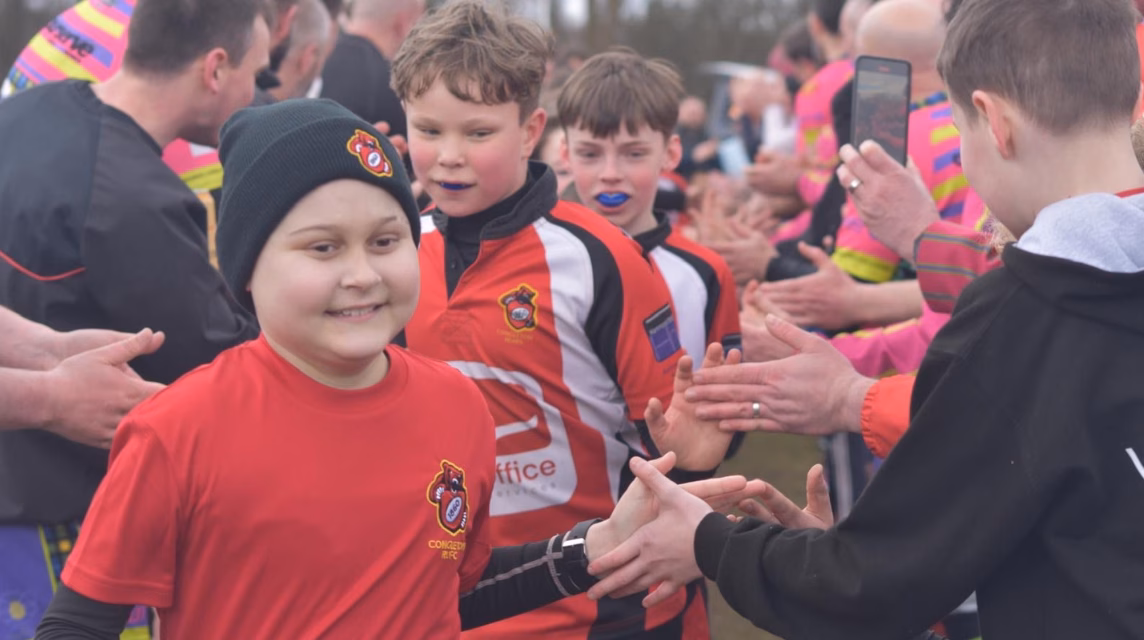 A young boy wearing a black hat and a red t-shirt. He is surrounded by other young boys about to play in a rugby match.