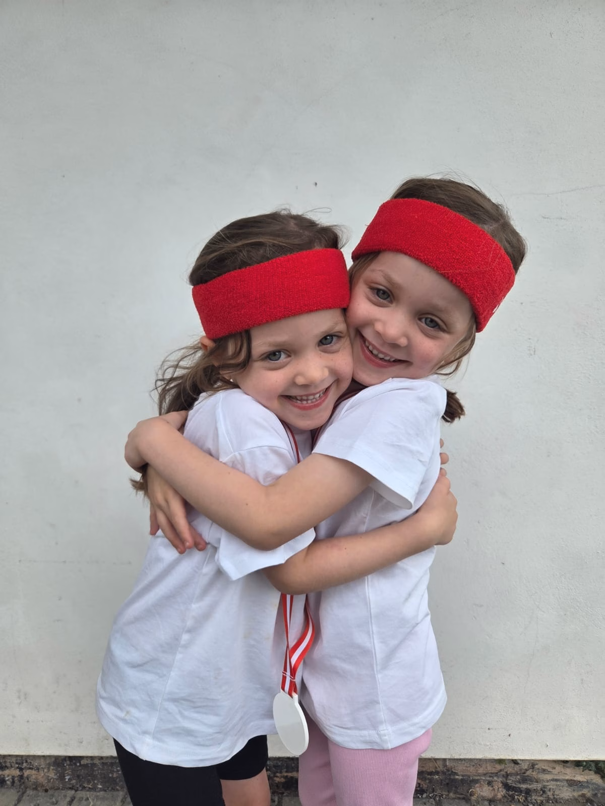 Two young girls hugging. They both wear red sweatbands on their heads and white t-shirts.