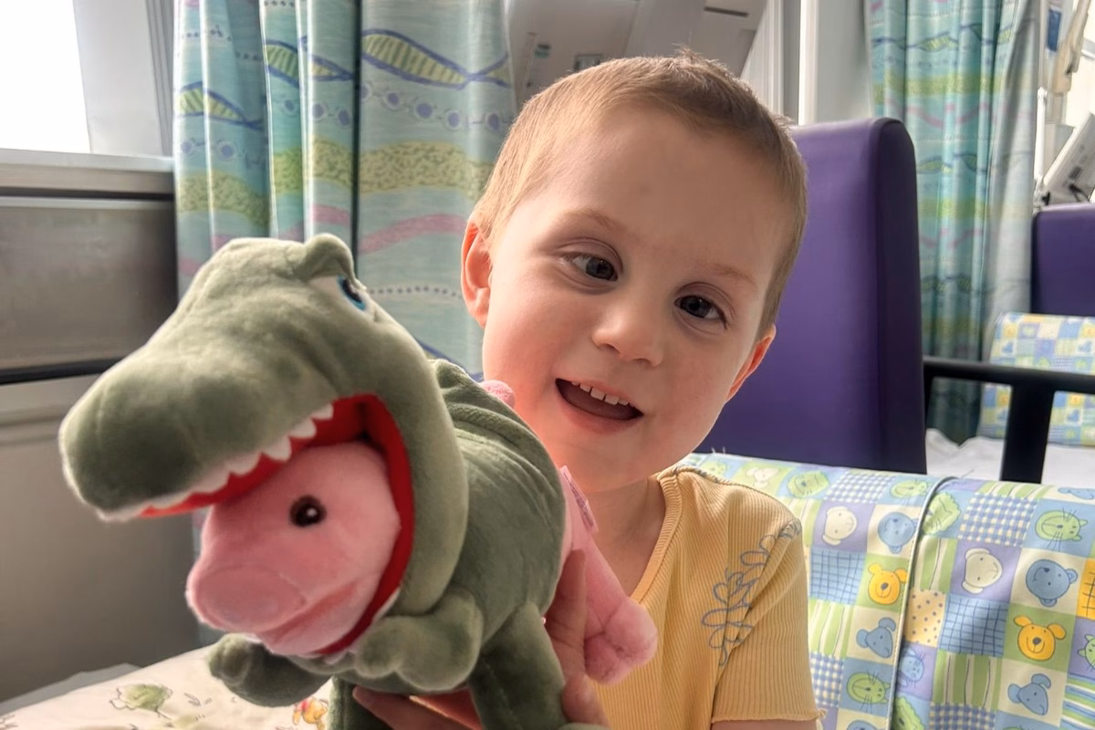Girl in hospital bed holding toy pig wearing dinosaur costume