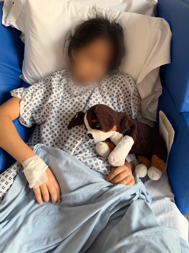 Girl in hospital bed, face blurred out, holding soft toy dog