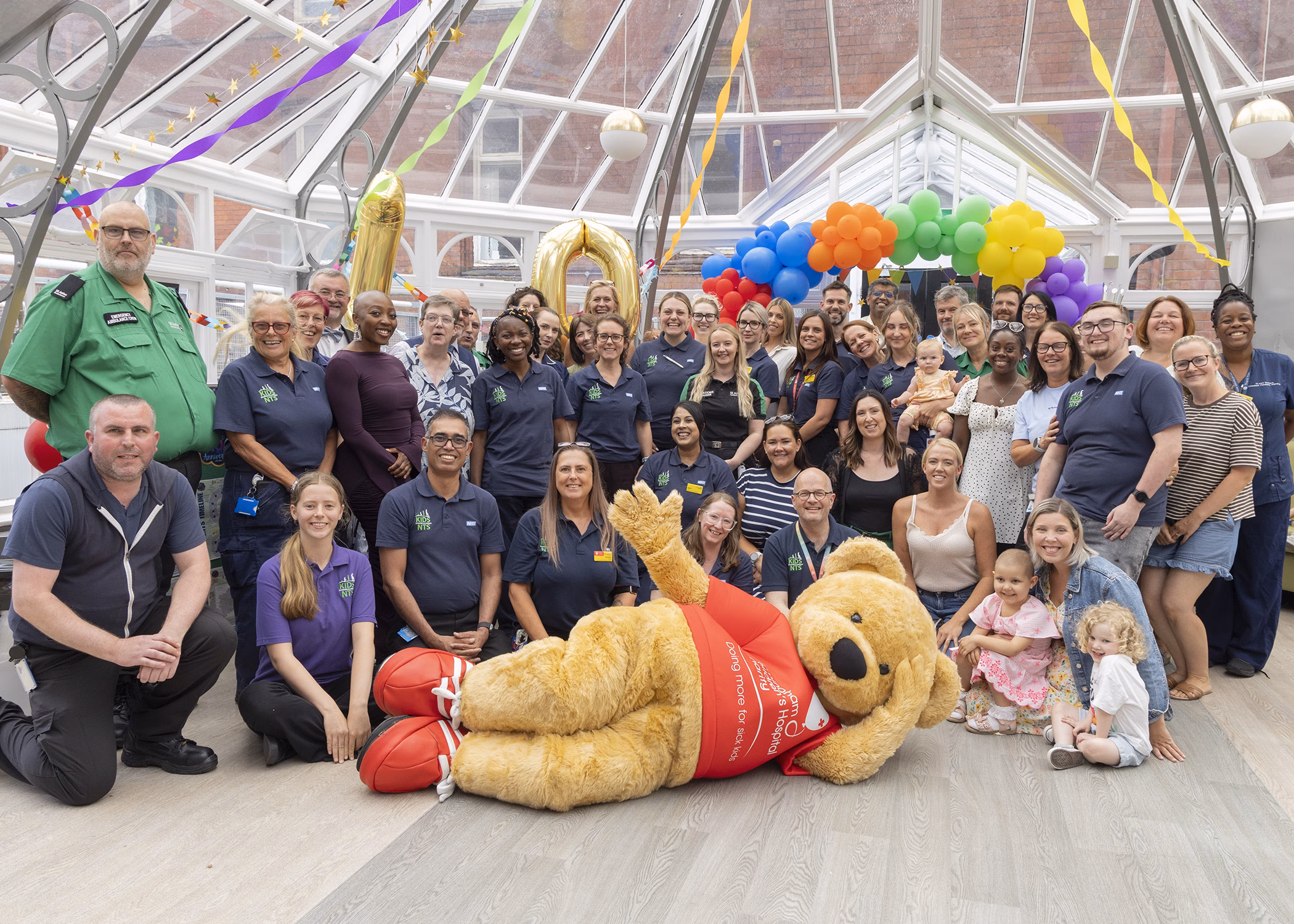 Smiling KIDS team at celebration with Bertie Bear Mascot lying down and posing