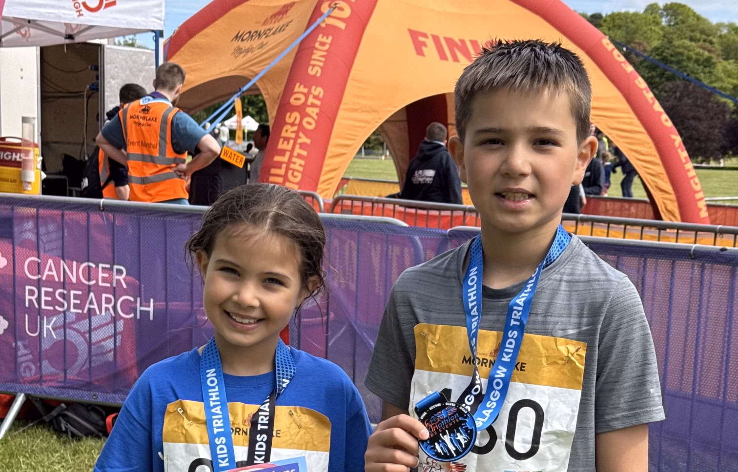 Girl and boy in running clothes wearing medals, standing in front of tent in field