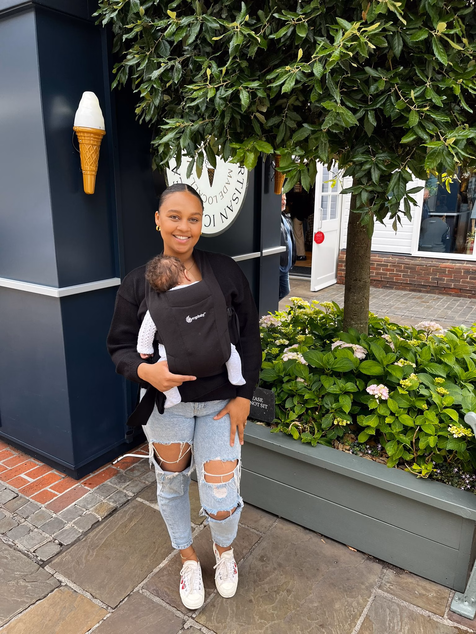 Woman standing outside ice cream parlour holding baby with baby carrier